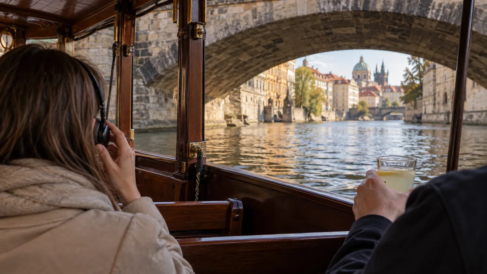 Guests inside a small wooden Prague canal boat, with a listener using headphones as the boat passes beneath a stone bridge.